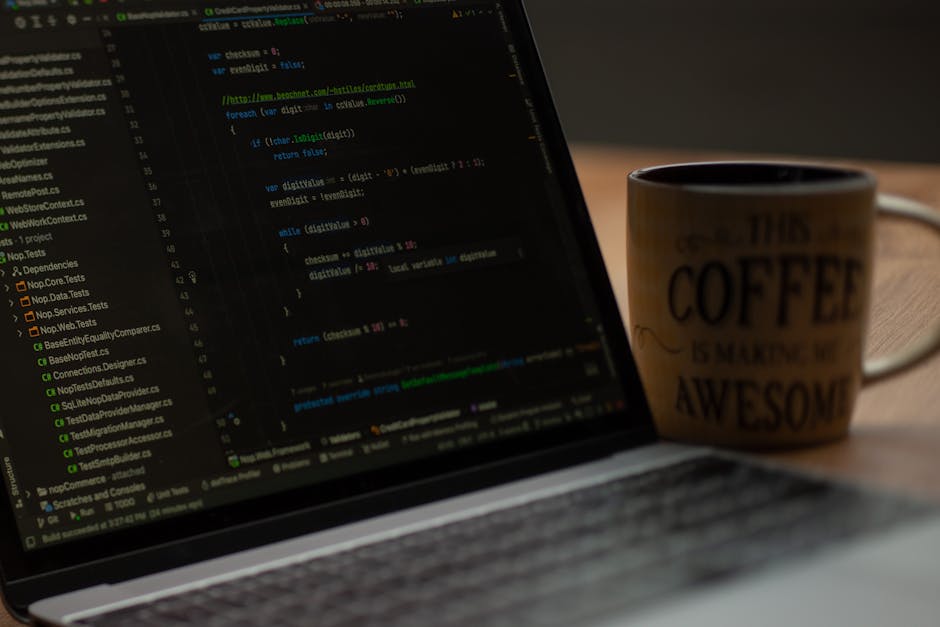 The image shows a person wearing a light blue shirt seated at a desk in a well-lit office environment. They are focused on a laptop displaying lines of colorful code, with their right hand holding a p